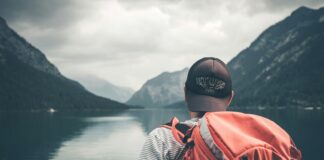 man with red hiking backpack facing body of water and mountains at daytime