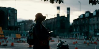 woman wearing coat standing on road with travel luggage during daytime