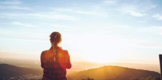 person sitting on wood fence facing mountain under cloudy sky