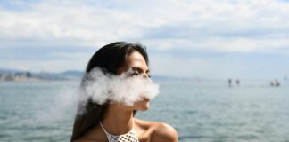 woman wearing white sleeveless top smoking tobacco while standing near blue sea under white and blue skies during daytime