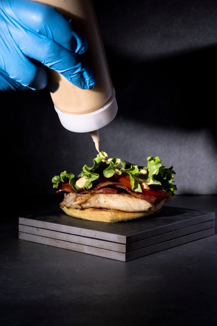 a person in blue gloves is putting ketchup on a sandwich