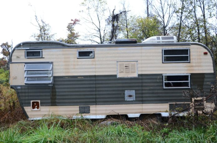 Old camper trailer sits abandoned in overgrown grass.